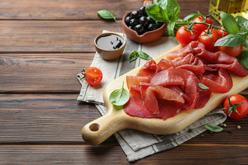 Board with delicious bresaola served with tomato and basil leaves on wooden table. Space for text