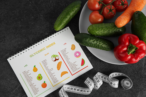 Notebook with information about glycemic index, measuring tape and vegetables on grey table, flat lay