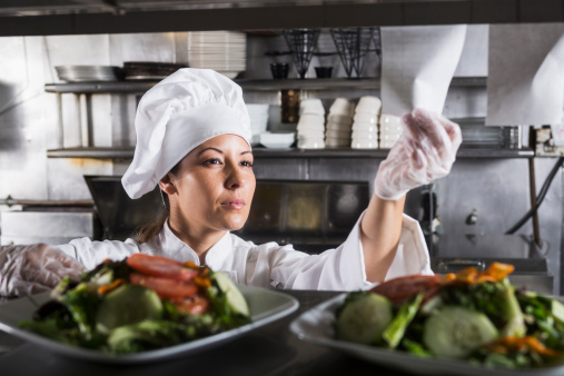 Female Hispanic chef (30s) in commercial kitchen, preparing gourmet salad.