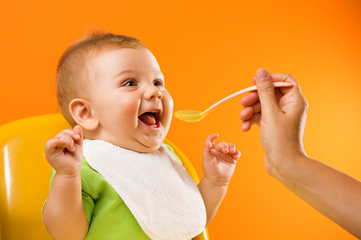 Mother's hand feeding an excited funny baby with bib in front of a vivid orange background.