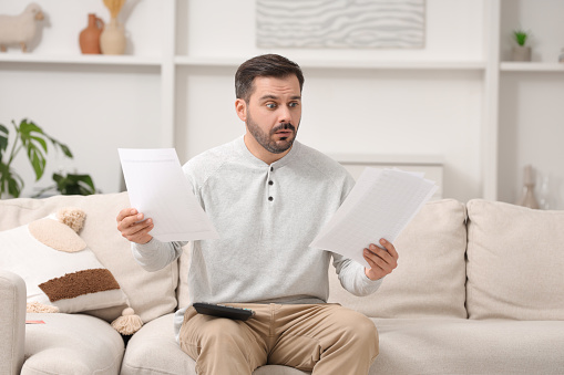 Shocked man with debt notifications and calculator planning budget at home. Financial problem