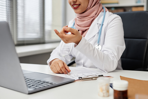 Cropped shot of female medical practitioner wearing hijab consulting patient remotely using laptop, focus on medical records