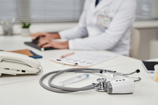 Medium shot with selective focus on stethoscope, digital and infrared thermometers, doctors hands typing on keyboard in blurred background