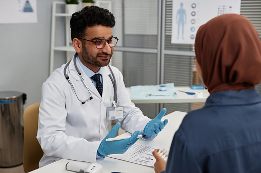 Medium shot of Middle Eastern man doctor talking to female patient in hijab about treatment at clinic