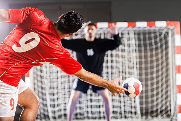 Handball player in red shooting toward a net Penalty shoot. Rear view of handball player shooting at goal, while goalkeeper is in the background with his hands raised.    handball stock pictures, royalty-free photos & images