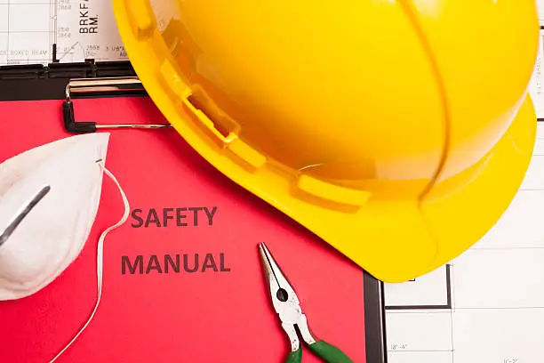 A safety manual is surrounded by work gloves, a hardhat, safety goggles and work tools.