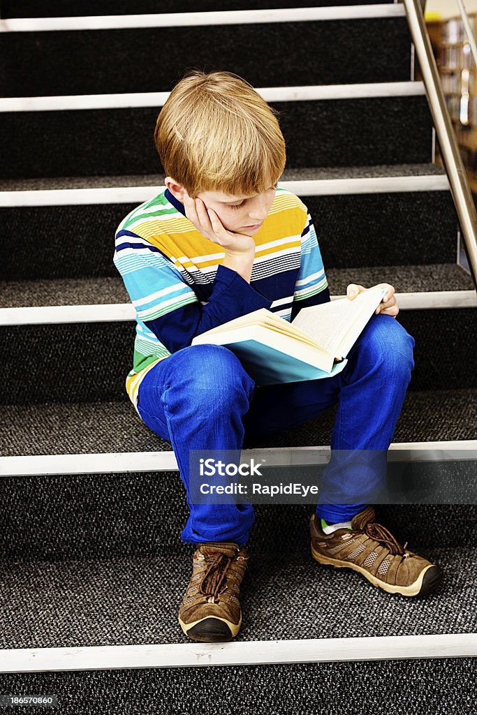 Totalmente ma perennemente capelli biondi ragazzo si trova la lettura in biblioteca passaggi - Foto stock royalty-free di 10-11 anni