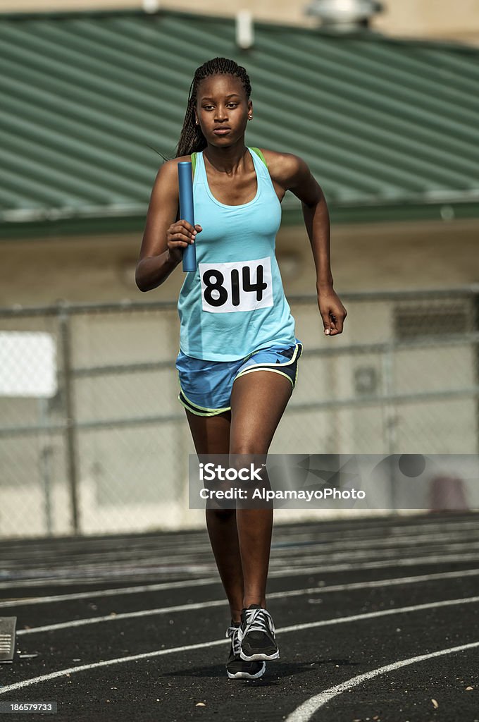 Young female track competitor running relay race - III High school female athlete running raley race with relay baton. Stadium Stock Photo