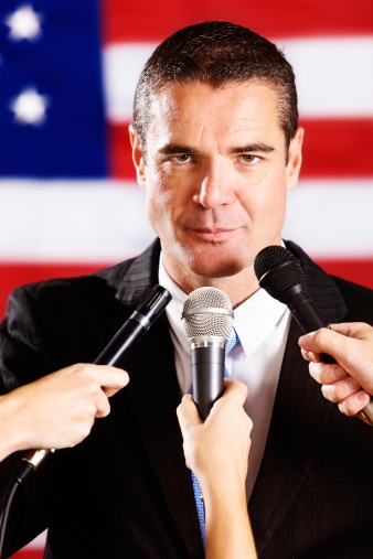 This handsome, confident-looking man, probably a candidate for political office, stands in front of the US flag, facing many microphones being held up eagerly by the Press.