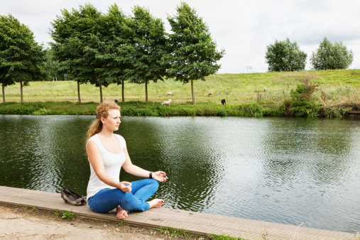 beautiful candid young woman with red hair sitting and meditating at river in summer