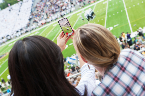 2 young women spectators taking a picture at sports event with a smart phone.