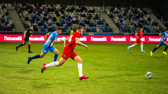 Male soccer players running and leading ball during match on sports field, side view wide shot