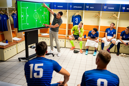Male soccer coach showing players positioning on large interactive screen, planning game strategy in locker room