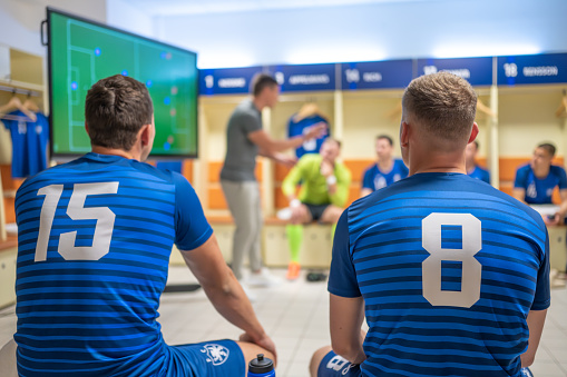 Male soccer players team listening to coach explaining game strategy in locker room before match, selective focus