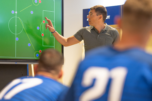 Male soccer coach showing players positioning on large interactive screen, planning game strategy in locker room