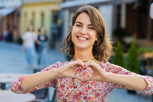 I love you. Caucasian woman makes symbol of love, showing heart sign to camera, express romantic feelings, express sincere positive feelings. Charity, gratitude donation. Outdoors in urban city street