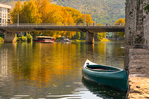 Golden autumn landscape in historical tourist city with bridge Trebinje Bosnia and Herzegovina snow-free cityscape, boat near ancient stone fortress, reflected in surface of river, yellow trees Balkan