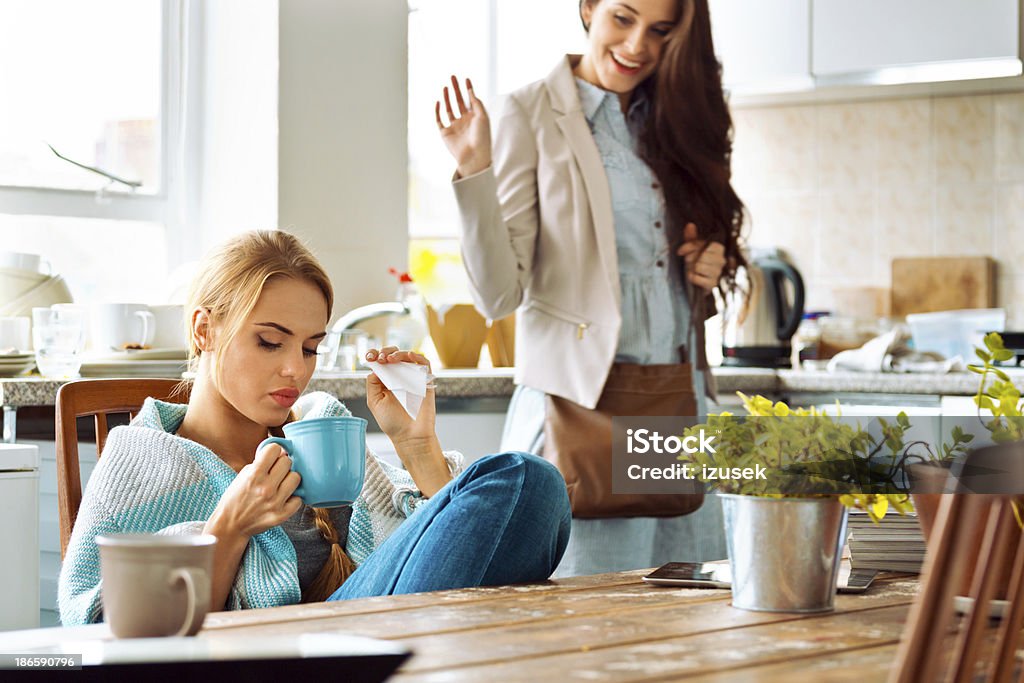 Students lifestyle Young woman has a cold, sitting in a kitchen wrapped in blanket, holding teacup and handkerchief, with her roommate going out and saying goodbye. 20-24 Years Stock Photo