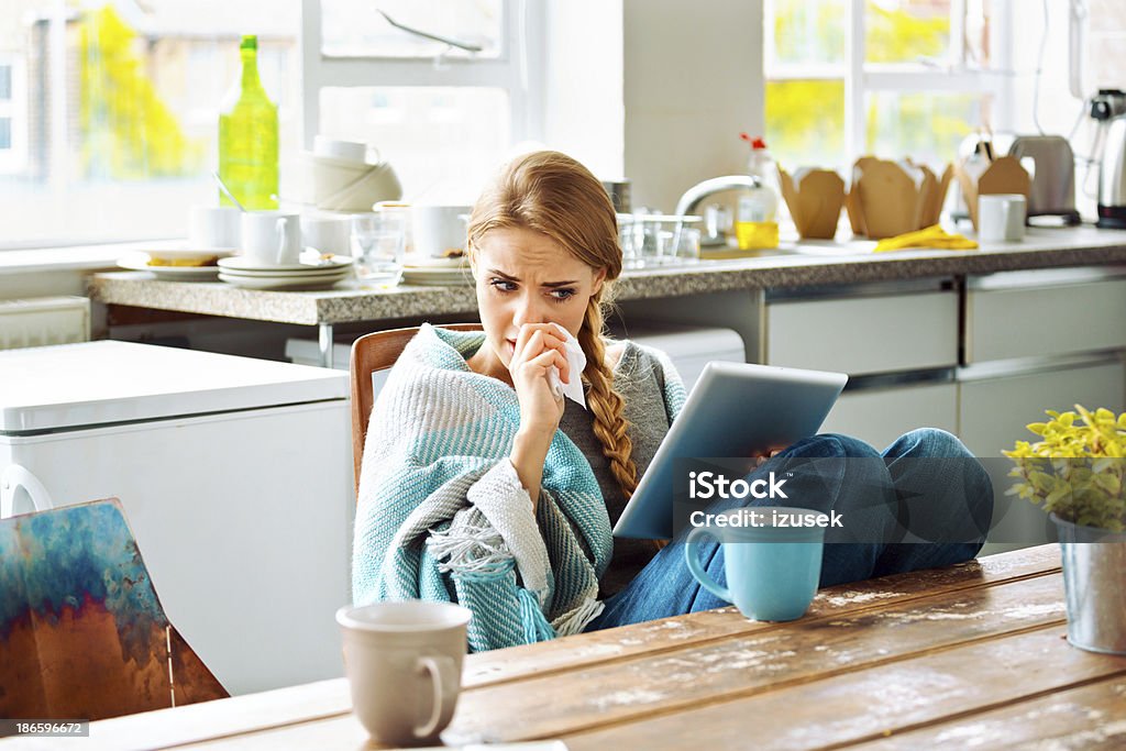 Sick woman Young woman has a cold, sitting in a kitchen wrapped in blanket and using a digital tablet. Cold And Flu Stock Photo