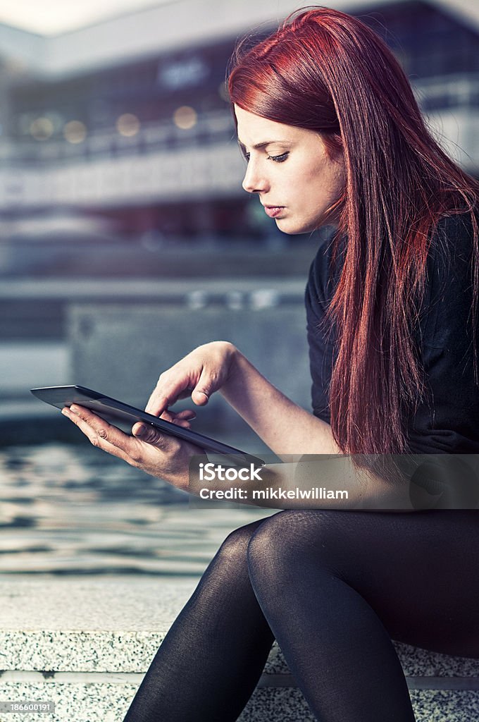 Young woman is surfing the web or playing a game Woman with red hair is sitting down. She is holding a digital tablet and touches the screen. 20-29 Years Stock Photo