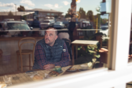 View from outside through a window looking in at a middle aged man with a disability who uses a wheelchair sitting at a table solo while enjoying coffee and pastries.
