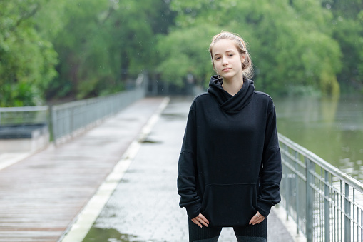 Portrait of young beautiful female blond hair crossed arms in black coach jacket standing in nature park looking at camera.
