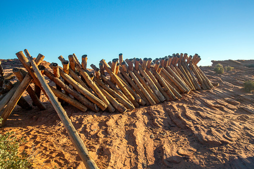 Stacks of wood arranged in the sand of the desert. The early morning sunlight casts stark shadows on the desert sands, with a pile of posts, used for the construction of an unfinished fence. The blue cloudless sky is a sharp contrast to the landscape below.