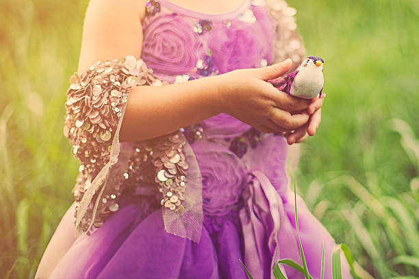 Portrait of a cute little two year old girl dressed in an adorable purple tutu dress and sparkly top, standing outside in nature. Details of the little girl holding onto a little bird.
