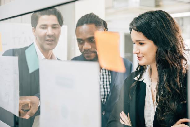 istock A diverse group of professionals meets in the office, using post-it notes on a glass board to brainstorm. They promote teamwork, leadership, and successful business discussions. 1866264939