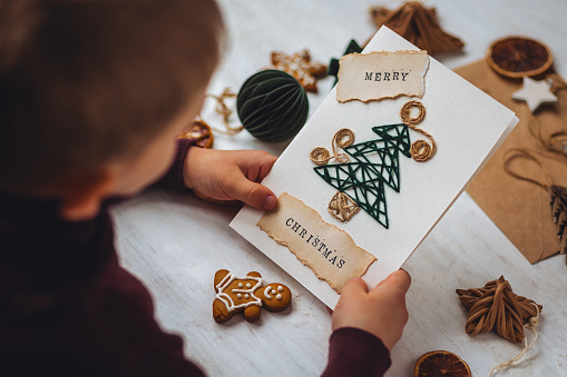 Concept of New year craft, kid's holiday leisure activity creating presents. Recyclable materials. Little boy holding handmade postcard with fir tree and printed message merry Christmas