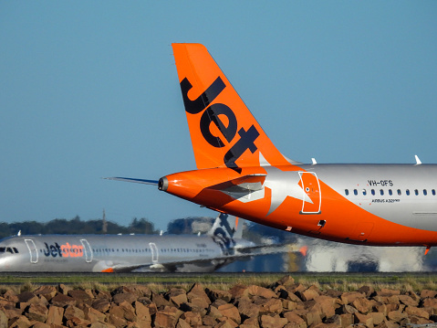 A Jetstar Airbus A321-251NX plane, registration VH-OFS, taxiing to the southern end of the main runway of Sydney Kingsford-Smith Airport for departure as flight JQ37 to Denpasar.  In the background is a Jetstar A321-231 plane, registration VH-VWU, taxiing after arrival from the Gold Coast as flight JQ419. This image was taken from Kyeemagh, Botany Bay, on a hot and sunny afternoon on 14 December 2023.