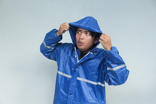 Young Asian man with worried expression about it's going to rain soon in blue raincoat on gray background. Rainy season concept.