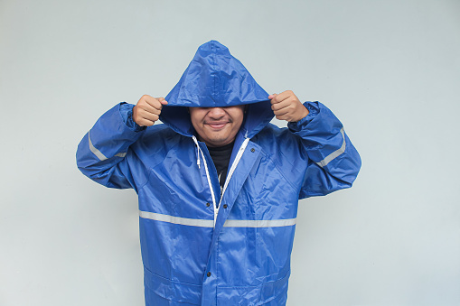 Asian young man with funny expression in blue raincoat on gray background. Rainy season concept.