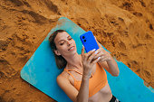 Fit woman in orange sports top enjoys a break during her workout to capture a selfie, embracing the digital connection amidst her outdoor fitness routine by the sandy lakeside