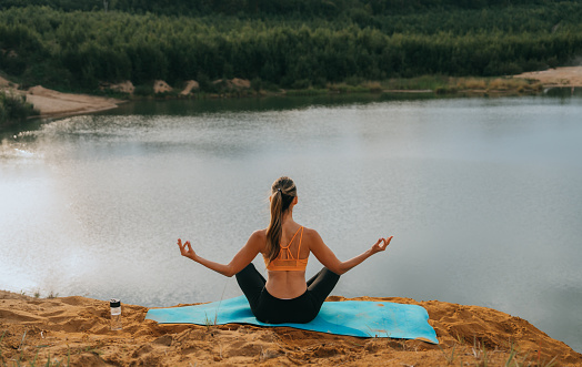 Rear view on fit girl in sportswear sitting on yoga mat in meditation faced to lake and forest. Healthy lifestyle, outdoor training, mental health. American female trainer relaxing on weekend.