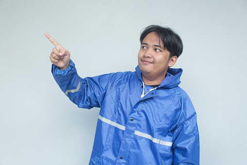 Young Asian man with happy expression in blue raincoat pointing at copy space beside him, isolated in gray background.