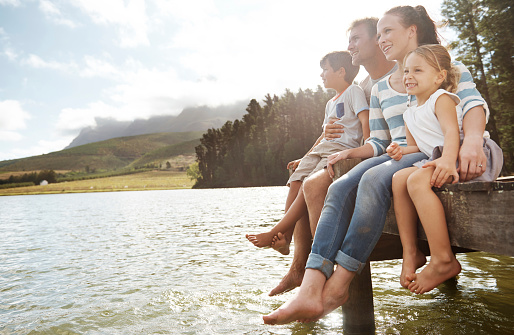 Side view of a smiling family of four sitting on the edge of a pier and admiring the lake