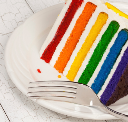 Slice of colourful rainbow layered birthday cake decorated with sprinkles and buttercream icing.