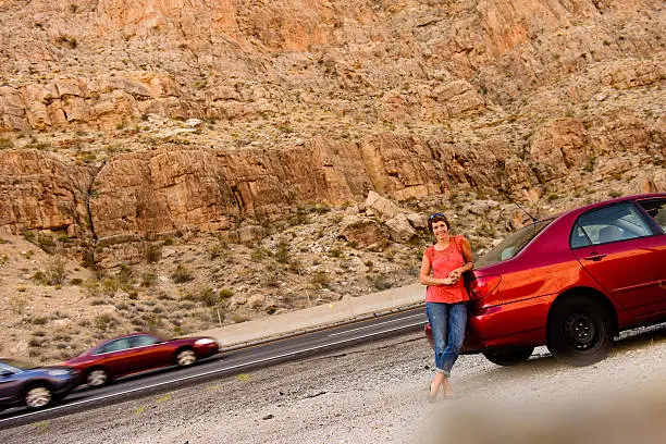 A woman on a road trip, leaning on her car.  Behind her, cars pass, and a red mountain from Southern Utah is in the background.