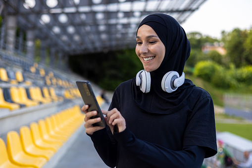 Muslim sportswoman in hijab at stadium with headphones and phone in hands, woman during active exercise and fitness, outdoors listening to online music from audio books and podcasts app.