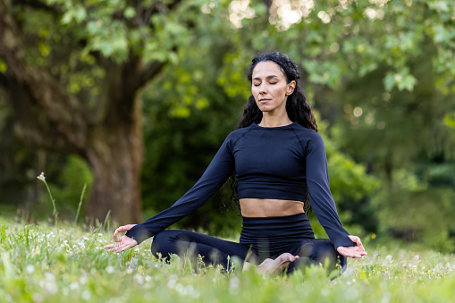 Young beautiful hispanic woman in park sitting on fitness mat and meditating in lotus pose, woman resting after fitness and running with eyes closed, breathing fresh air in tracksuit.