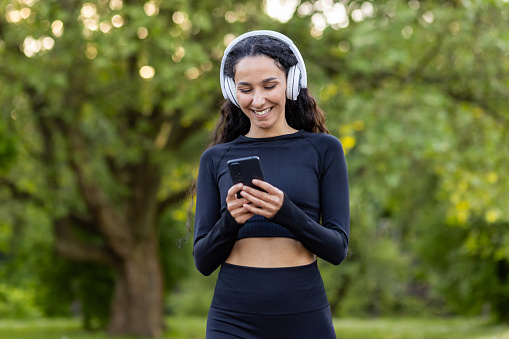Successful slim latin american woman in park during active exercise and jogging uses smartphone app, sportswoman smiling in headphones listening to audio podcasts and online radio.