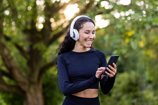 Young beautiful Latin American woman walks in the park during active physical exercises, woman uses an application for listening to audio books and music on phone, smiling sportswoman in headphones.