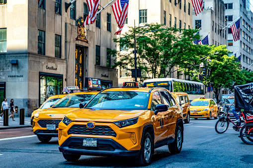 New York, USA; May 31, 2023: The typical Big Apple yellow cab, driving down Fifth Avenue in the middle of Manhattan with the American flags in the background.
