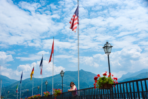 Woman standing next to a railing with several national flags in the village of Menaggio, Lake Como, Italy.