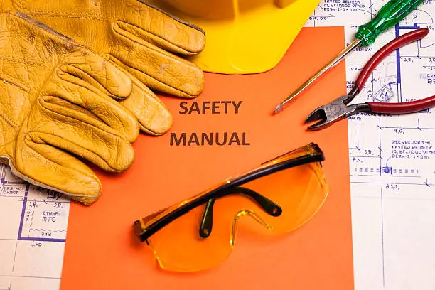 A safety manual is surrounded by work gloves, a hardhat, safety goggles, work tools, and floor plans.