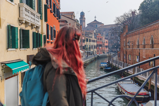 Young woman with dyed red hair and backpack standing on a pedestrian bridge over a canal, visiting Venice, Italy