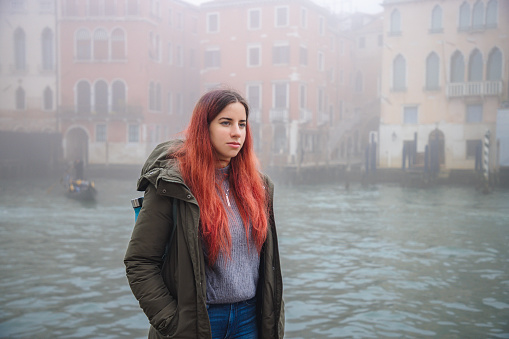Beautiful young woman with long dyed red hair standing by the Grand Canal on a foggy winter day, traveling to Venice, Italy