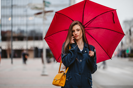 Portrait of a beautiful young woman with a red umbrella talking on her smart phone on the city street in autumn.