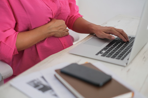 Close-up shot of unrecognizable pregnant professional working from home. She's sitting in front of an open laptop at her desk, typing on the keyboard. She put one hand on her big belly.